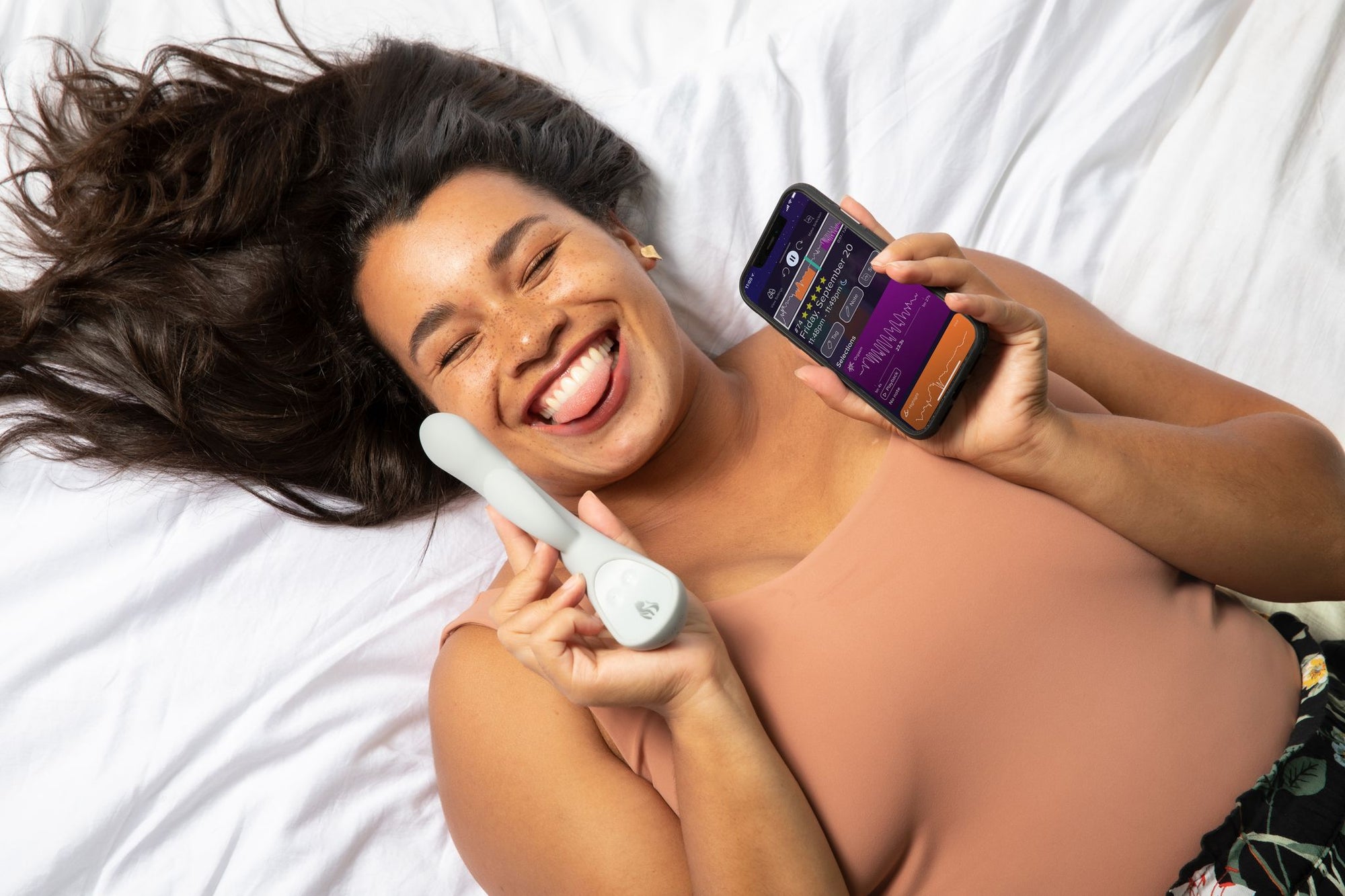 Mujer tumbada en una cama y sacando la lengua mientras sostiene el vibrador inteligente Lioness y la aplicación